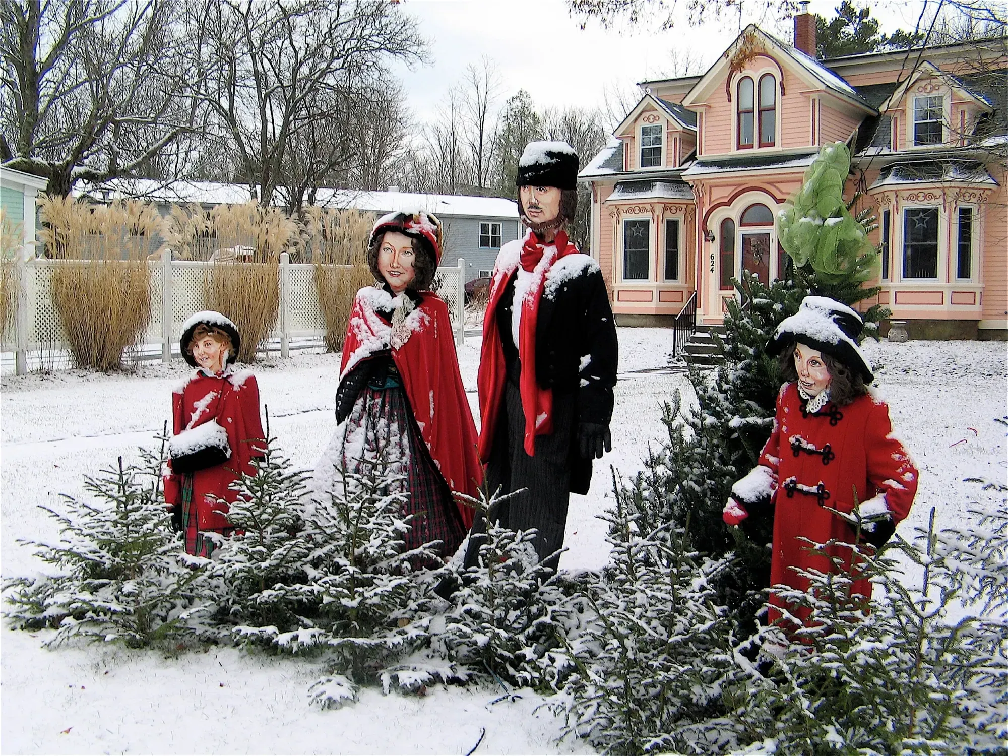Handmade Victorian caroller figures in red cloaks stand in a snowy front yard before a pink heritage house during the Season of Father Christmas