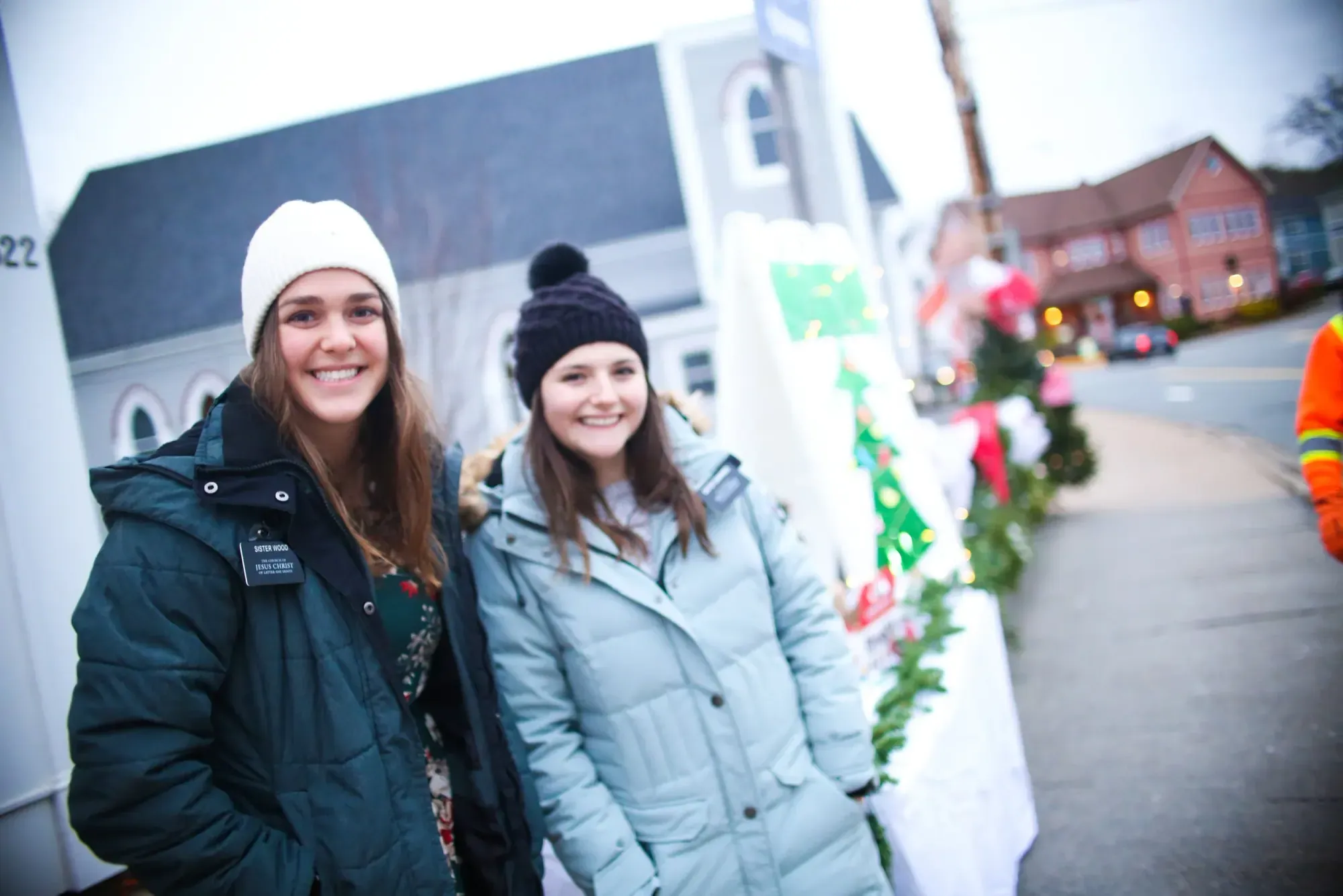 Two bundled-up festival-goers smile on a decorated street at dusk during the Season of Father Christmas in Mahone Bay