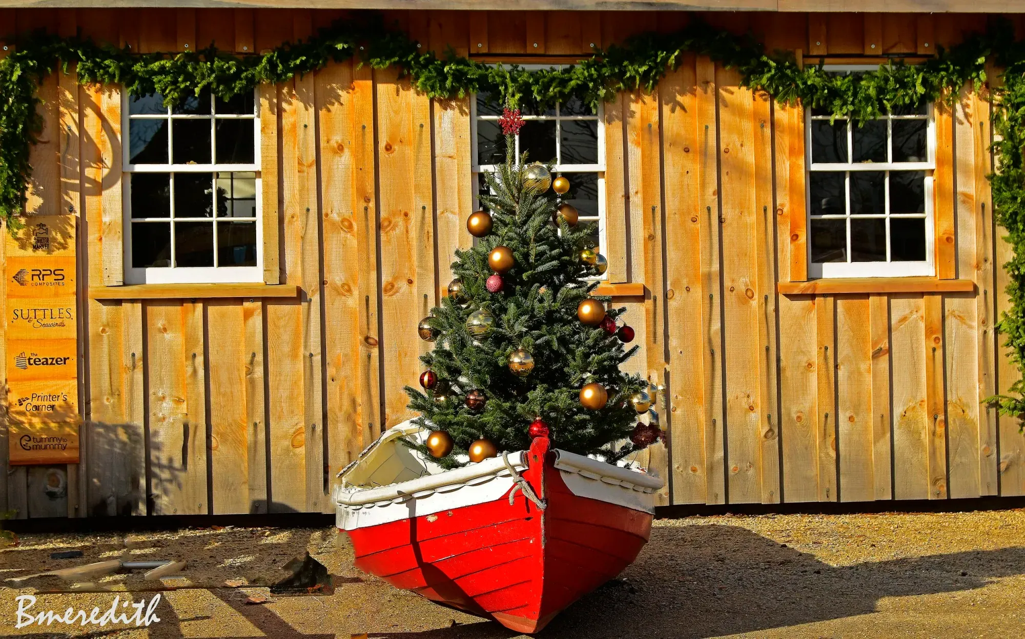 A decorated Christmas tree stands in a red-and-white wooden dory against a garland-trimmed building — a classic seaside Mahone Bay Christmas