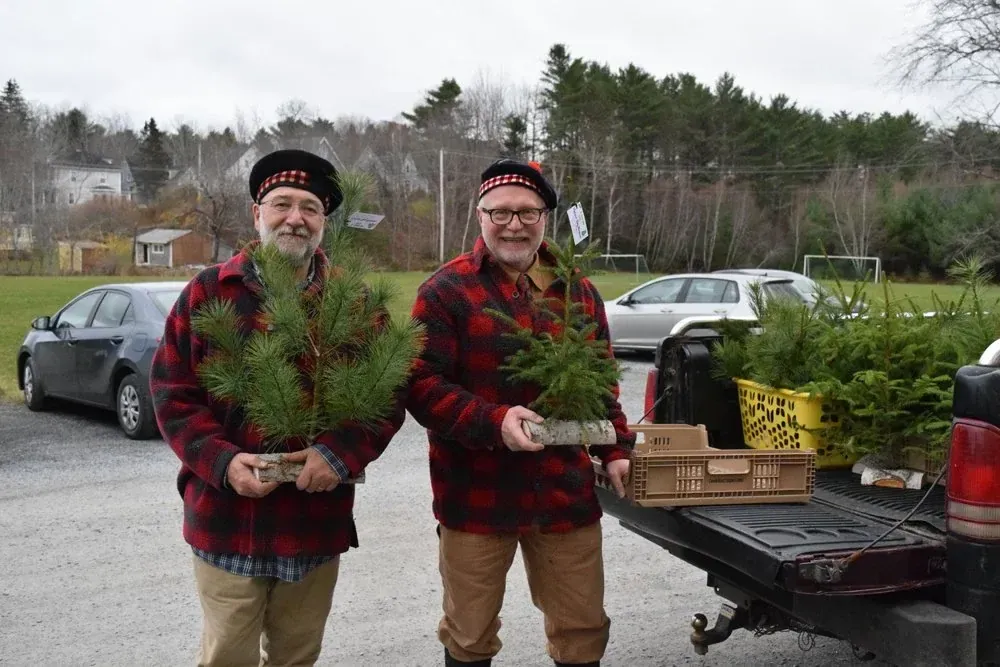 Two volunteers in tartan caps hold balsam fir trees beside a loaded truck while preparing for the Season of Father Christmas
