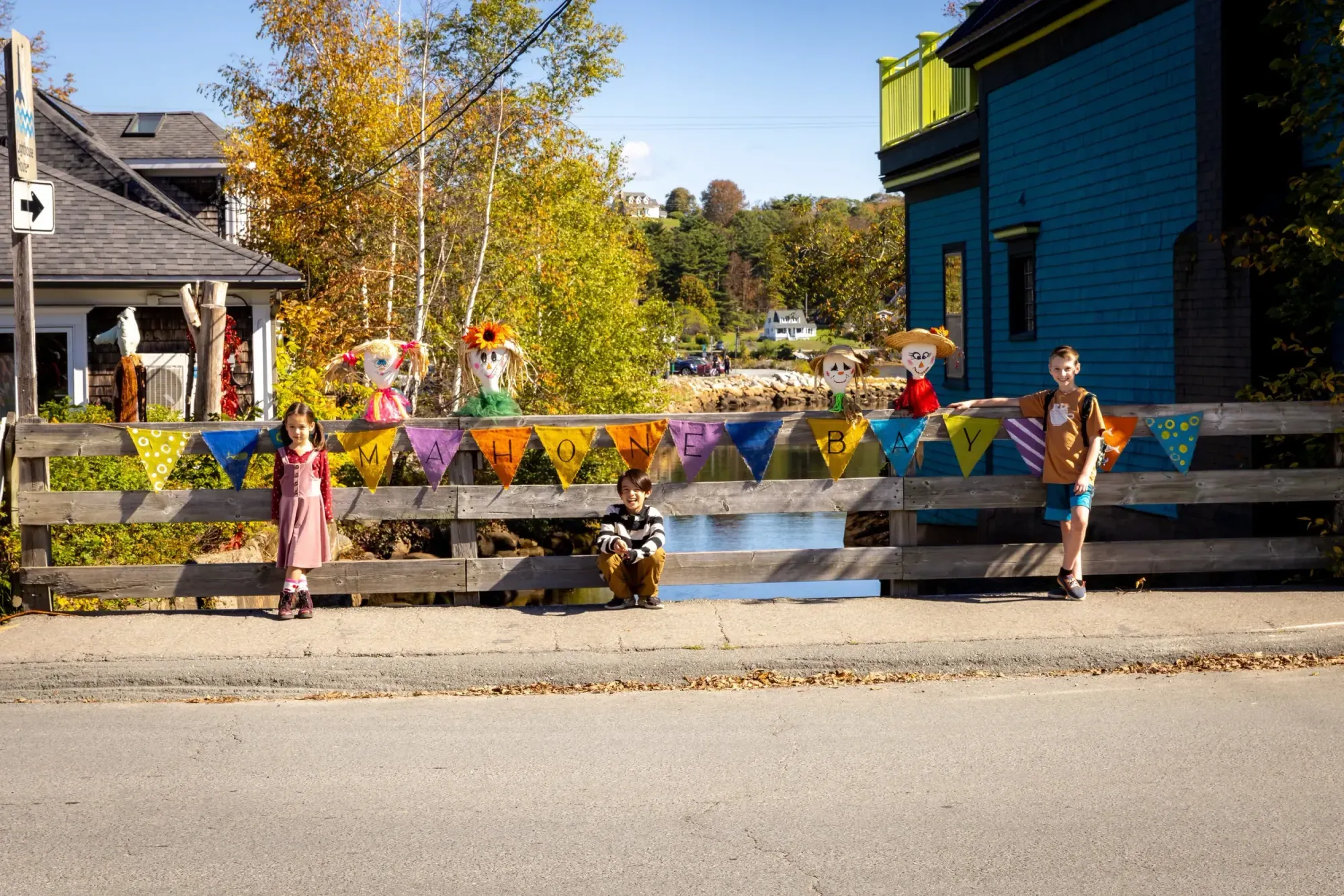 Children and handmade scarecrows beside a MAHONE BAY pennant banner on the town's waterfront during the Scarecrow Festival